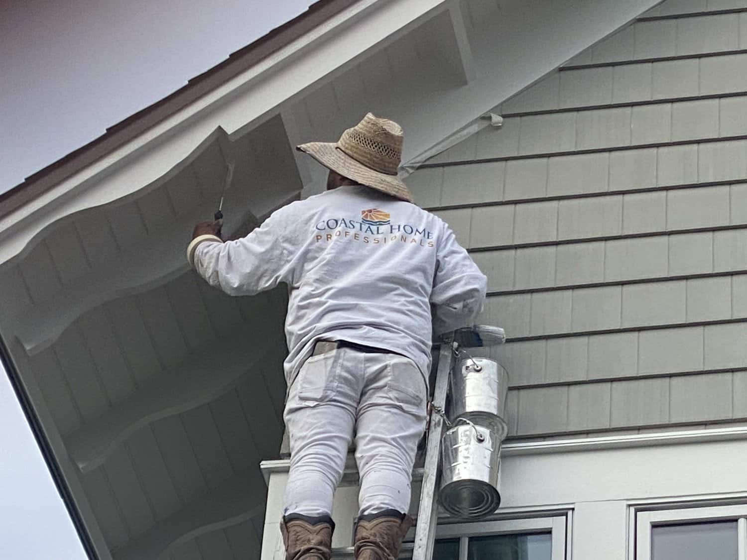 A Coastal Home Professional painter is on a ladder and painting the eaves of a roof
